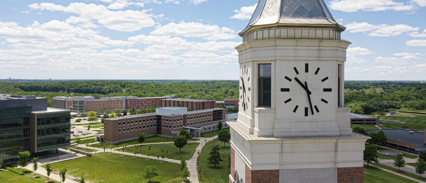 A photo of the clock face of Elliott Tower with Oakland University's campus in the background.